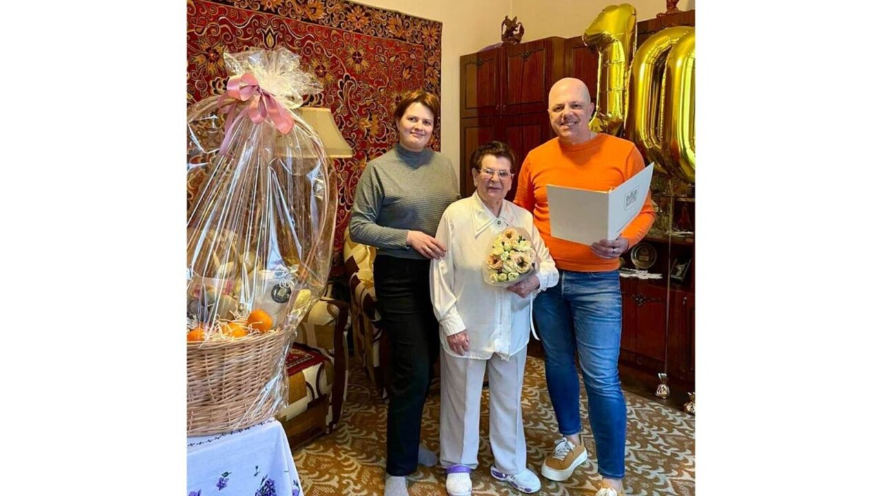 Ганна Сопрунова (по центру) з букетом квітів, позаду видно золоті повітряні кульки з цифрою “100”. Ліворуч на столі стоїть великий подарунковий кошик, а інтер’єр кімнати з килимом на стіні та дерев’яними меблями створює затишну, родинну атмосферу свята.